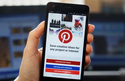 Image of a hand holding a cell phone that is displaying the Pinterest homepage.